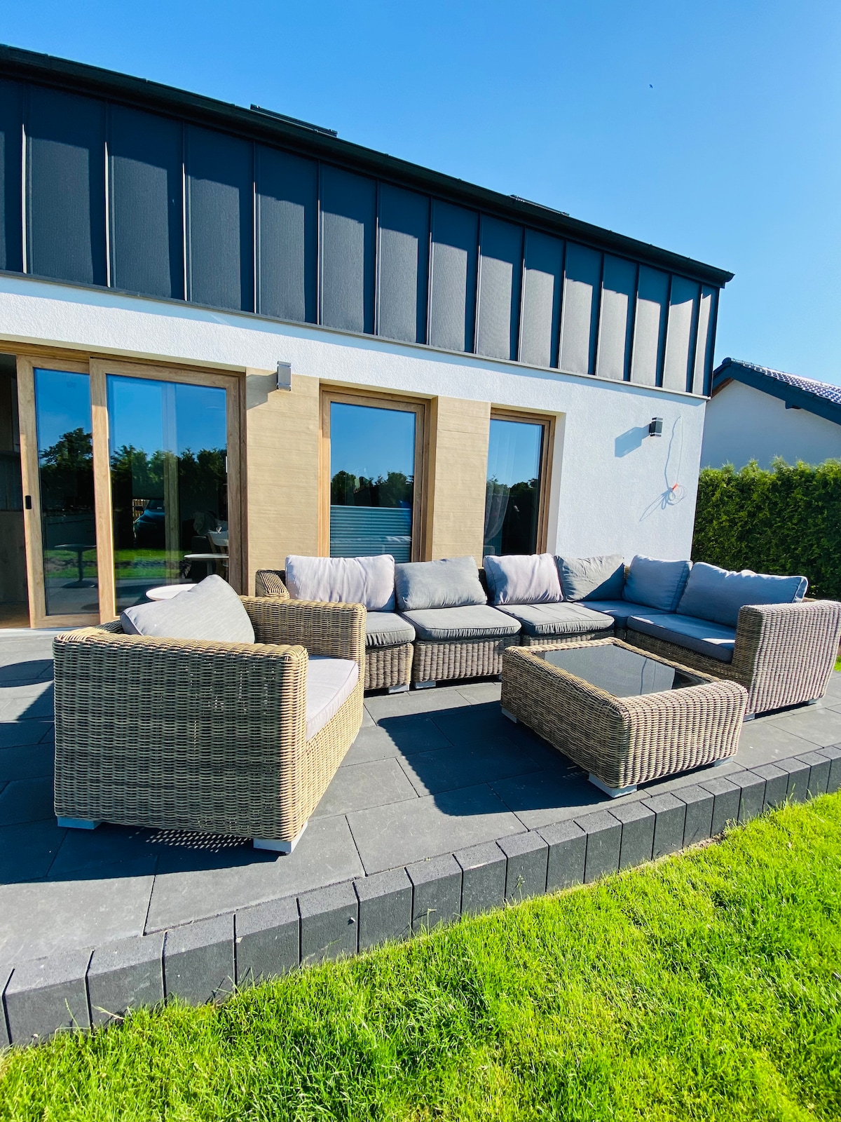 An outdoor seating area features a spacious arrangement of woven furniture, complemented by light gray cushions. A low table sits in the center, providing a comfortable space for relaxation, with sliding glass doors in the background allowing for seamless access to the indoor space.