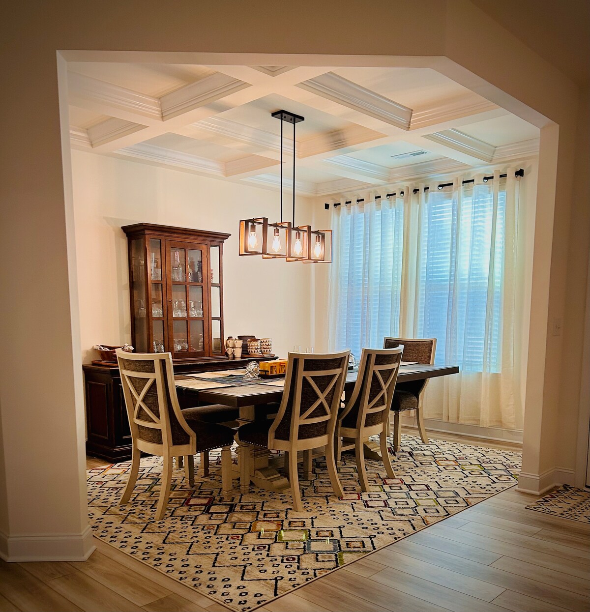 An elegantly appointed dining area features a large table surrounded by six chairs. A stylish cabinet showcases decorative items. Soft light filters through sheer curtains, creating a warm ambiance. The patterned rug complements the room's design, enhancing the overall atmosphere of the space.