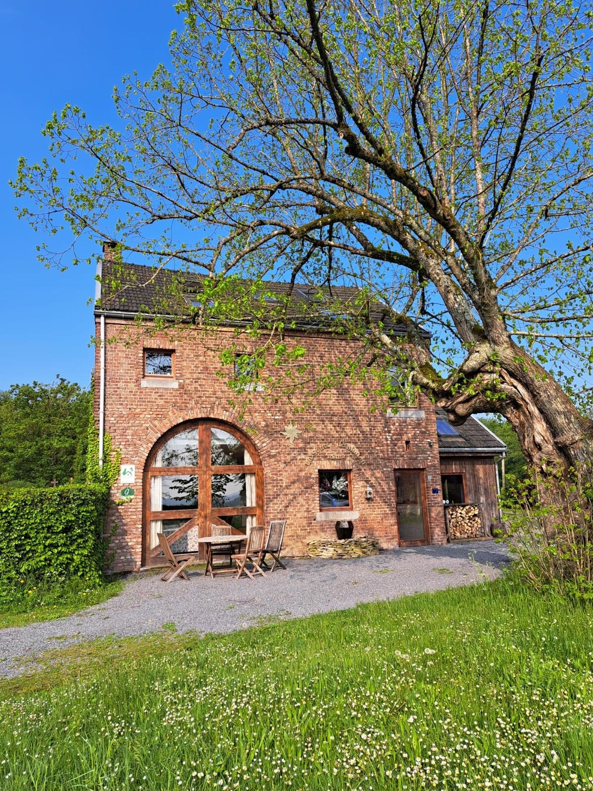 The exterior of a charming brick building is shown, featuring large arched windows and a wooden door. A small terrace with wooden furniture is positioned near a lush green area, complemented by a large tree. The setting exudes a peaceful, natural environment.
