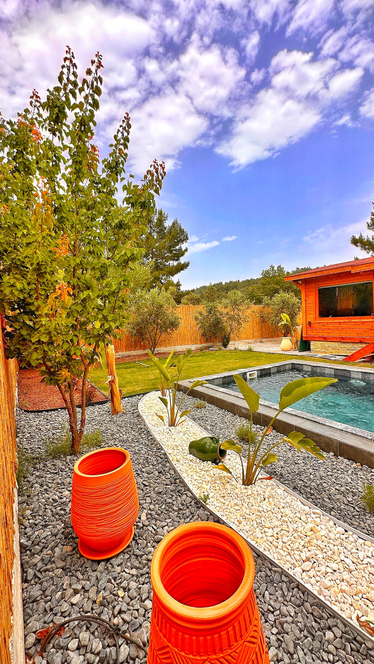 A vibrant outdoor space features two orange ceramic pots positioned on gravel, surrounded by lush greenery. A clear pool reflects the sky, while a wooden structure is visible in the background, completing a serene garden setting.