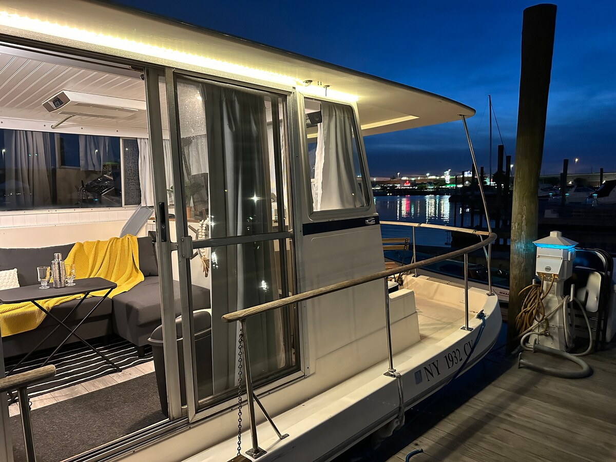 The boathouse is illuminated at night, showcasing large glass doors that provide a view into the living area. A table sits on the deck, inviting outdoor dining. The surrounding marina lights reflect on the water, enhancing the waterfront ambiance.