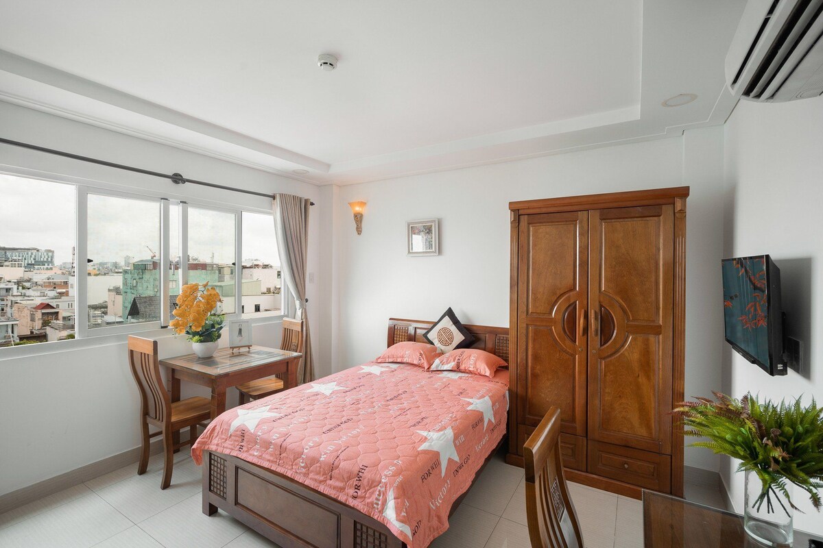 A well-lit room features a spacious bed with coral bedding and a wooden wardrobe. A small dining table with matching chairs is positioned near a large window, which offers a view of the surroundings. A flat-screen TV is mounted on the wall opposite the bed.