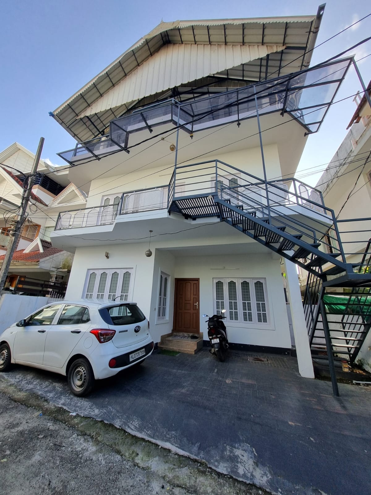 A three-story building features a modern façade with a metal staircase leading to the upper floors. Large windows allow abundant light into the structure. A driveway includes a parked car and a motorcycle, indicating accessibility and convenience.