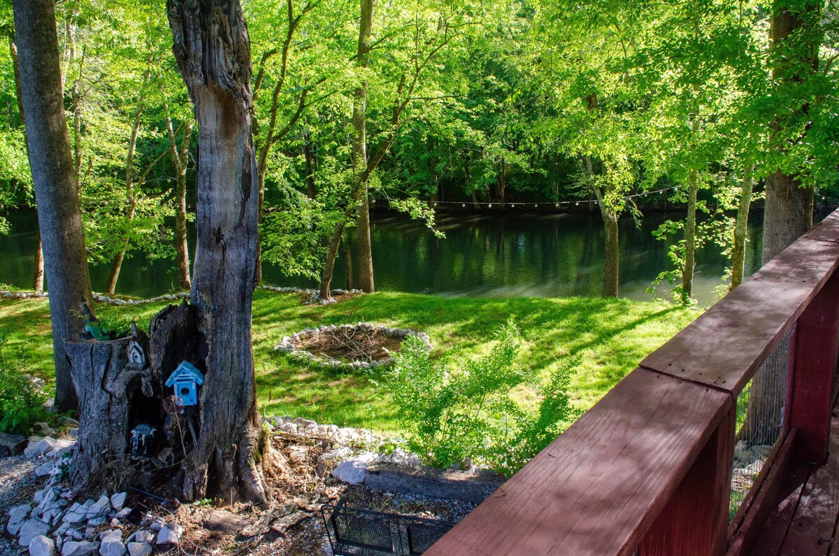 A tranquil outdoor scene showcases the Flint River surrounded by lush greenery. A fire pit is visible in the grass, complemented by a decorative birdhouse attached to a tree. Natural light filters through the leaves, enhancing the serene atmosphere.