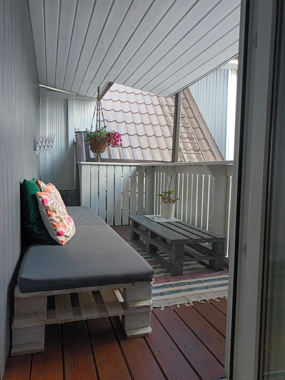 A private balcony is featured, furnished with a cushioned bench and a rustic pallet table. Potted flowers hang from the wall, adding a touch of color. Natural wood flooring and a striped rug contribute to a cozy ambiance, complemented by the sloped roof overhead.