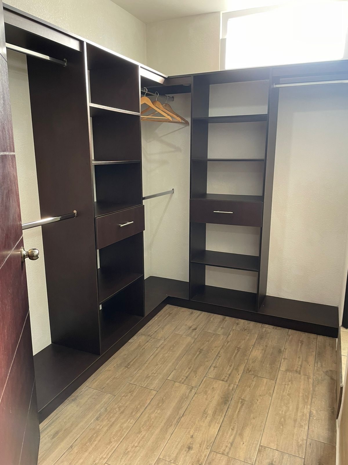 A spacious closet features dark wood cabinetry with multiple shelves, hanging space, and drawers. Natural light streams in from a small window, illuminating the organized storage area, which provides ample space for clothing and personal items.