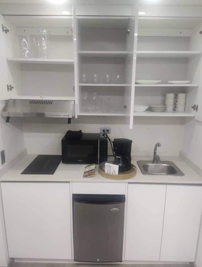The kitchenette features white cabinetry with upper shelves holding glassware and dishes. A compact coffee maker and microwave are present on the countertop, alongside a small sink. A refrigerator with a metallic finish is located below the counter, contributing to a functional and tidy cooking space.