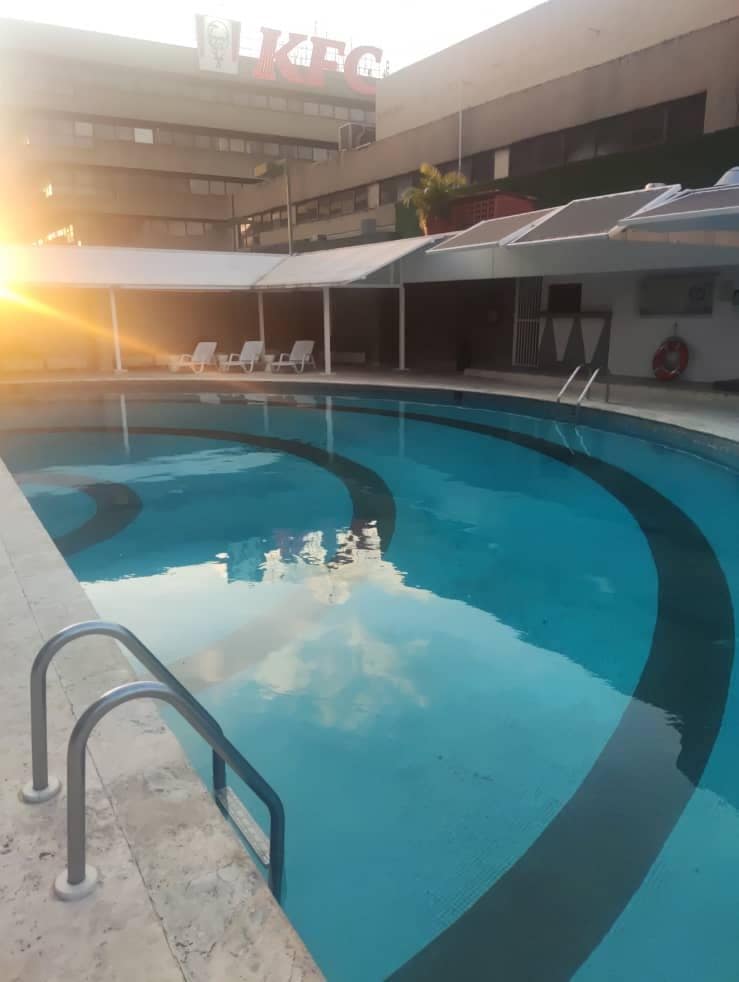 A glistening swimming pool is situated under a covered area, with gently sloping steps leading into the water. White lounge chairs are positioned around the pool, reflecting a calm atmosphere created by the surrounding structures and the soft light of the setting sun.