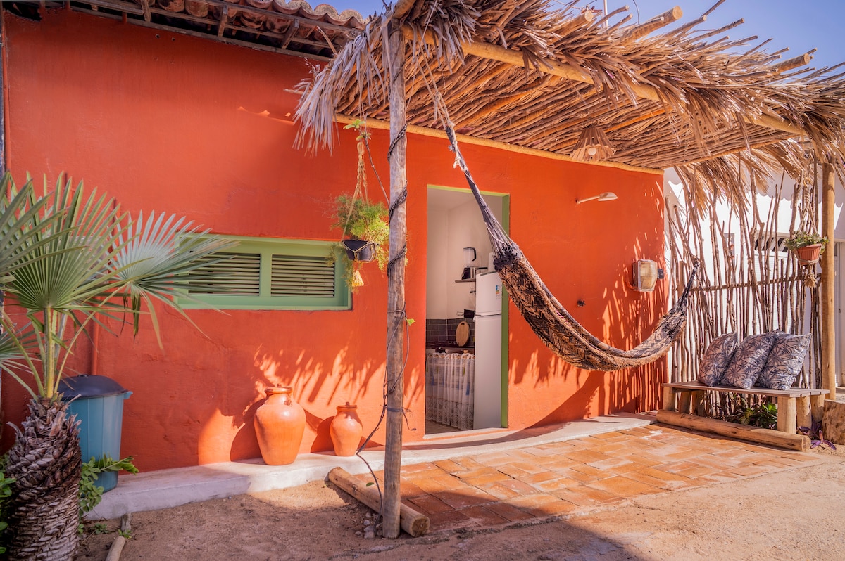 An inviting outdoor space features a vibrant orange wall, complemented by a straw roof. A wooden bench with decorative cushions provides seating, while a hammock is suspended nearby. Traditional clay pots and a palm plant add to the rustic charm of the area.