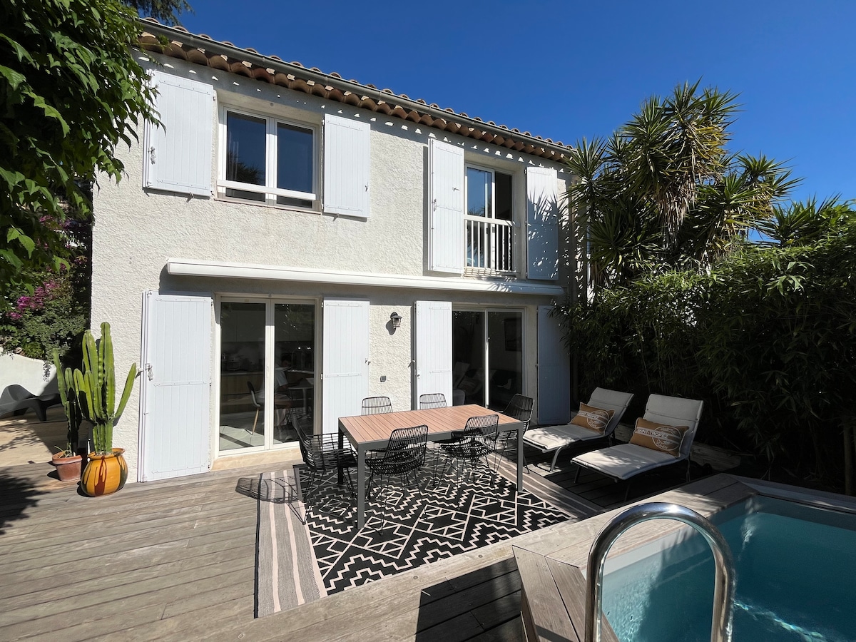 A serene outdoor area features a wooden deck with a dining table surrounded by six chairs, complemented by two lounge chairs. Lush greenery provides privacy. The modern house displays white shutters, large glass doors, and a balcony on the upper level, enhancing the inviting atmosphere.