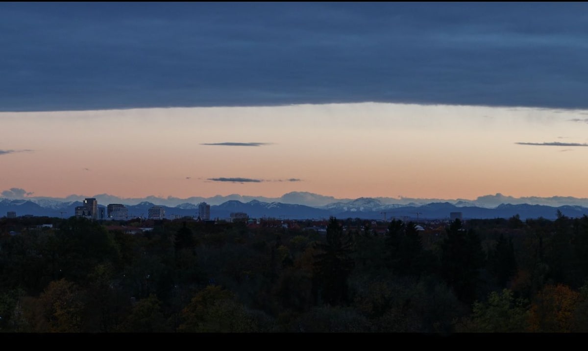 A panoramic view of the Munich skyline transitions into the snow-capped peaks of the Alps at dusk. Soft hues of orange and blue fill the sky, while the distant city structures and surrounding trees are silhouetted against the fading light.