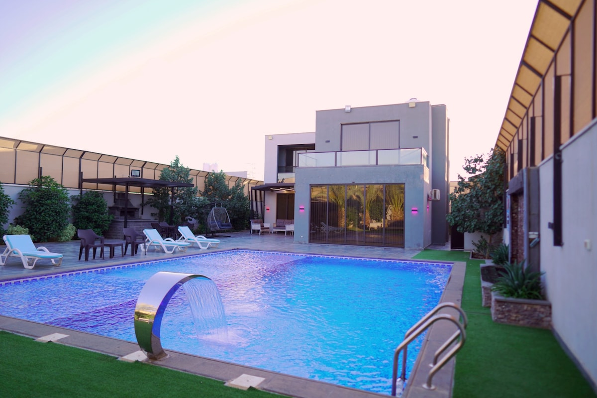 A spacious outdoor area features a large private pool with a gently cascading water feature. Lounge chairs are arranged around the pool, and a modern building with large windows stands in the background, providing an inviting space for relaxation and enjoyment.