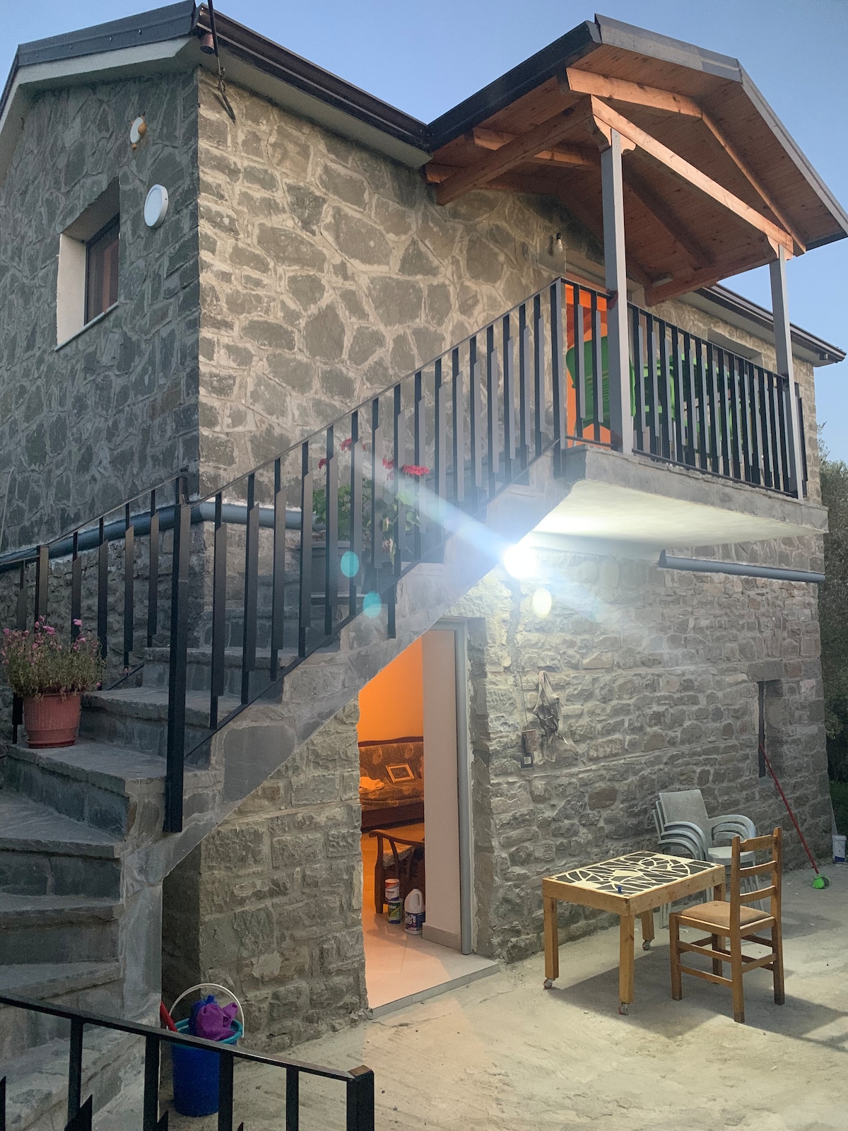 An exterior view of a two-story stone villa is presented, featuring a staircase leading to an upper balcony. The entrance is lit softly, revealing a glimpse of the interior. Nearby, a table and chairs are visible on the patio, surrounded by potted plants.