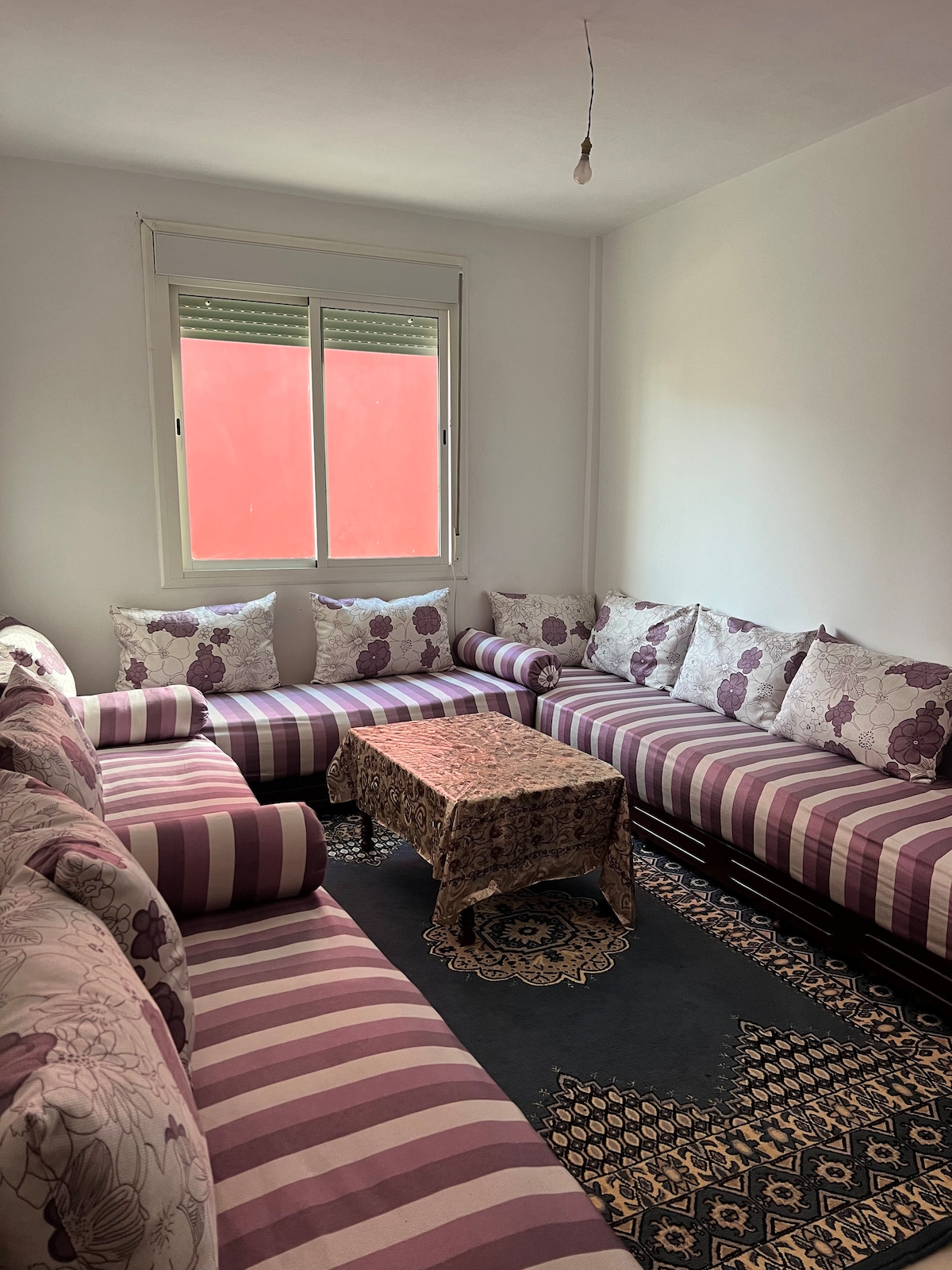 A comfortable sitting area is furnished with a large L-shaped sofa adorned with floral-patterned cushions. A low table is placed at the center, complemented by a decorative tablecloth. Light enters through a window, while a bright wall serves as a backdrop.