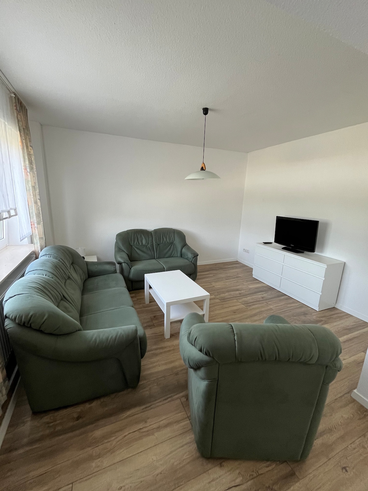 A well-appointed living area features two green sofas and two matching armchairs surrounding a central white coffee table. A media console stands against the wall, complemented by a wall-mounted television. Natural light fills the room through a large window adorned with light drapery.
