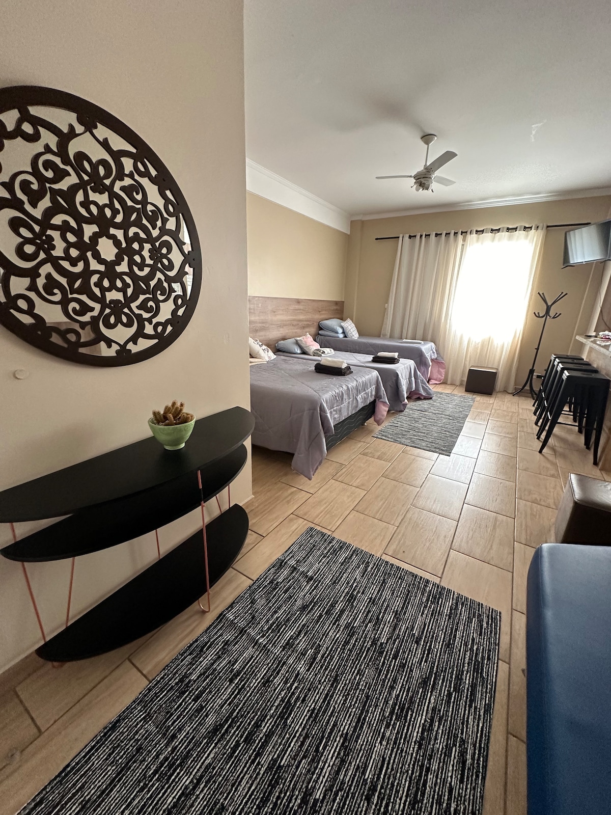 A spacious studio layout is featured, with two single beds and a double bed, all adorned with muted bedding. Natural light floods the area through a large window, while decorative elements, including a wall mirror and a small table with a plant, enhance the space.