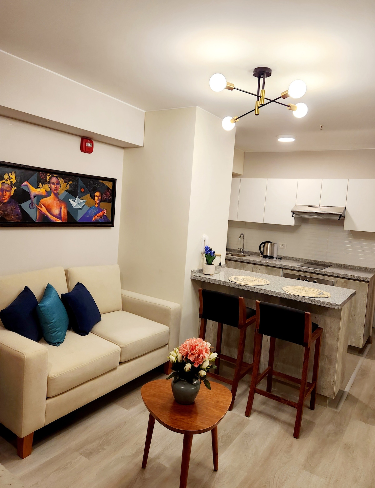 A cozy living space features a beige sofa adorned with blue cushions, creating a comfortable seating area. A wooden table with a floral centerpiece is positioned in front of the sofa. The kitchenette, equipped with modern appliances, lies in the background under stylish overhead lighting.