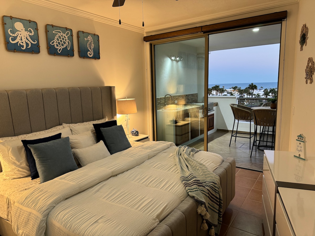A cozy bedroom is illuminated by soft light from a nearby window overlooking the sea. The spacious bed features a neutral-colored headboard and is adorned with blue decorative pillows. A light throw lies casually across the bed. A balcony with bar seating is visible through sliding glass doors.
