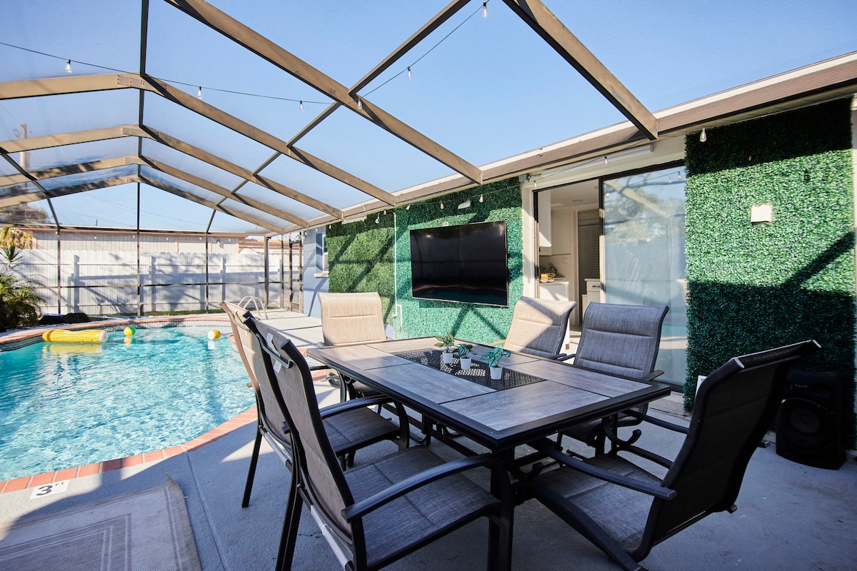 A shaded outdoor area features a large table surrounded by comfortable chairs, overlooking a well-maintained pool. A flat-screen TV is mounted on a green wall, and natural light brightens the space through the screened enclosure.