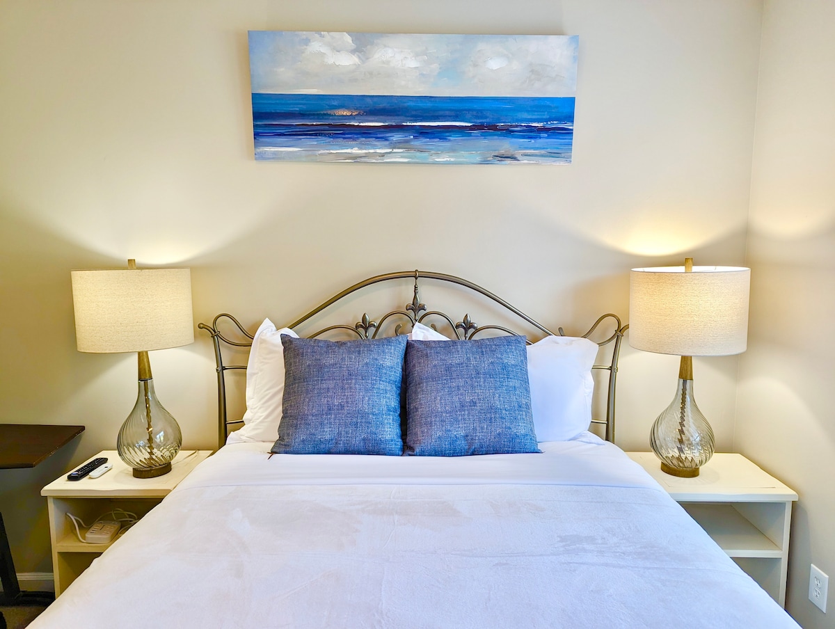 A cozy queen bed is positioned centrally in the image, adorned with two decorative blue pillows. Flanking the bed are two illuminated bedside lamps, providing ambient light. Above the headboard, a painting of a serene ocean landscape adds a touch of color to the room.