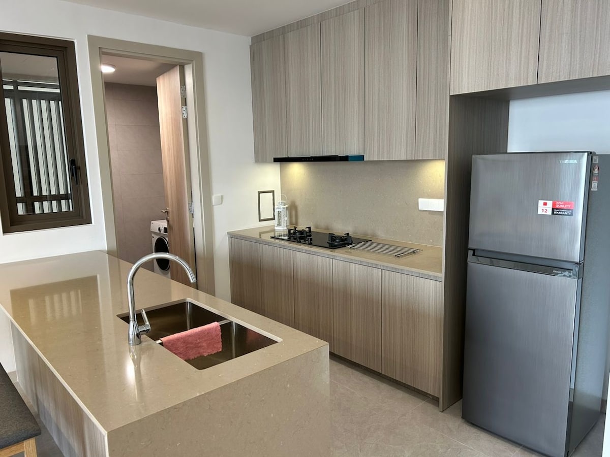 An open kitchen area is depicted, featuring light wooden cabinetry and a sleek countertop. A modern gas stove is positioned for cooking, while a stylish refrigerator stands nearby. Natural light enters through a window, enhancing the space's bright and functional design.