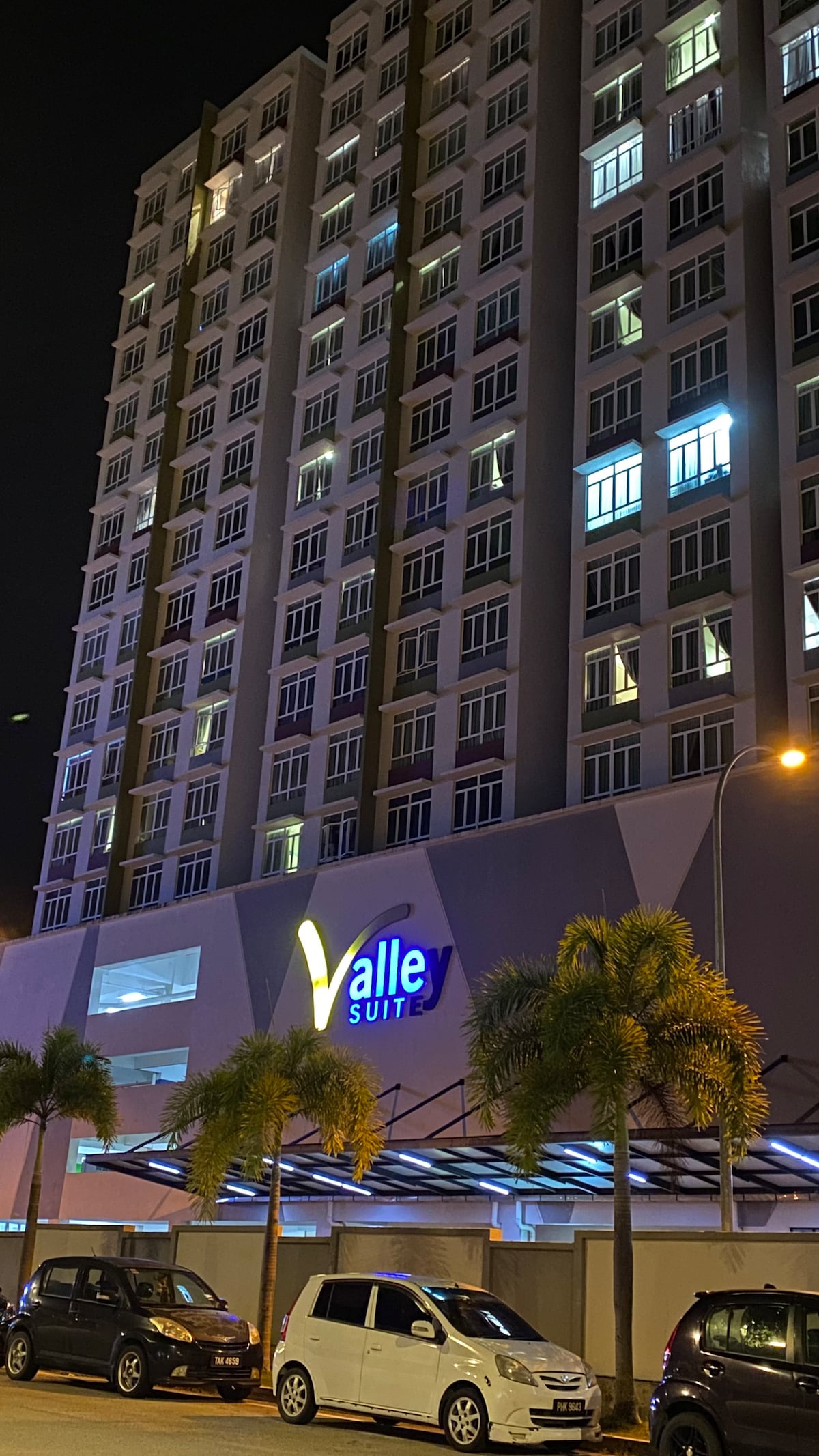 The exterior of the Valley Suite building is displayed at night, illuminated by soft lighting. The modern structure consists of multiple floors with large windows. A parking facility is visible in the foreground, alongside palm trees adding a touch of greenery.