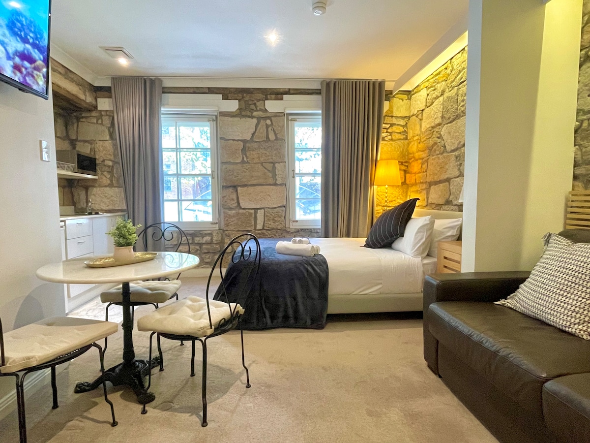 An inviting studio apartment is highlighted by historic sandstone walls and natural light from the windows. A spacious queen bed sits beside a leather sofa bed, while a small round dining table is positioned near the kitchenette. Soft fabrics and neutral tones create a relaxing environment.