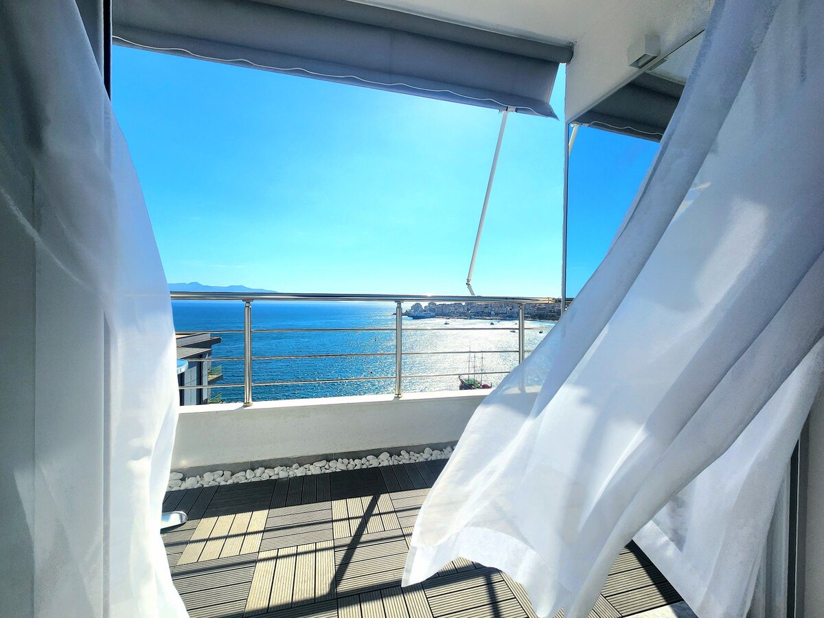 A view from a balcony framed by light curtains, revealing a bright blue sky and sparkling sea. The water reflects sunlight, creating a serene and open atmosphere. The balcony features a wooden deck that adds warmth to the setting.
