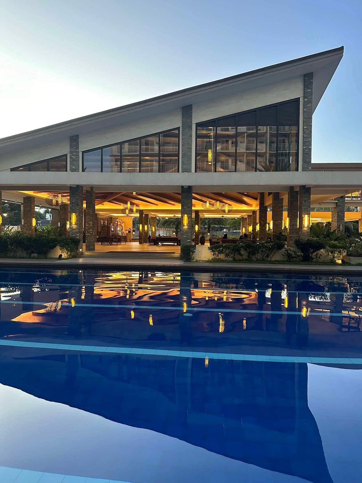 A modern architectural structure features a sloped roof and expansive glass windows, reflecting golden light from within. A clear pool in the foreground mirrors the building's design, while surrounding landscaping adds greenery to the serene setting.