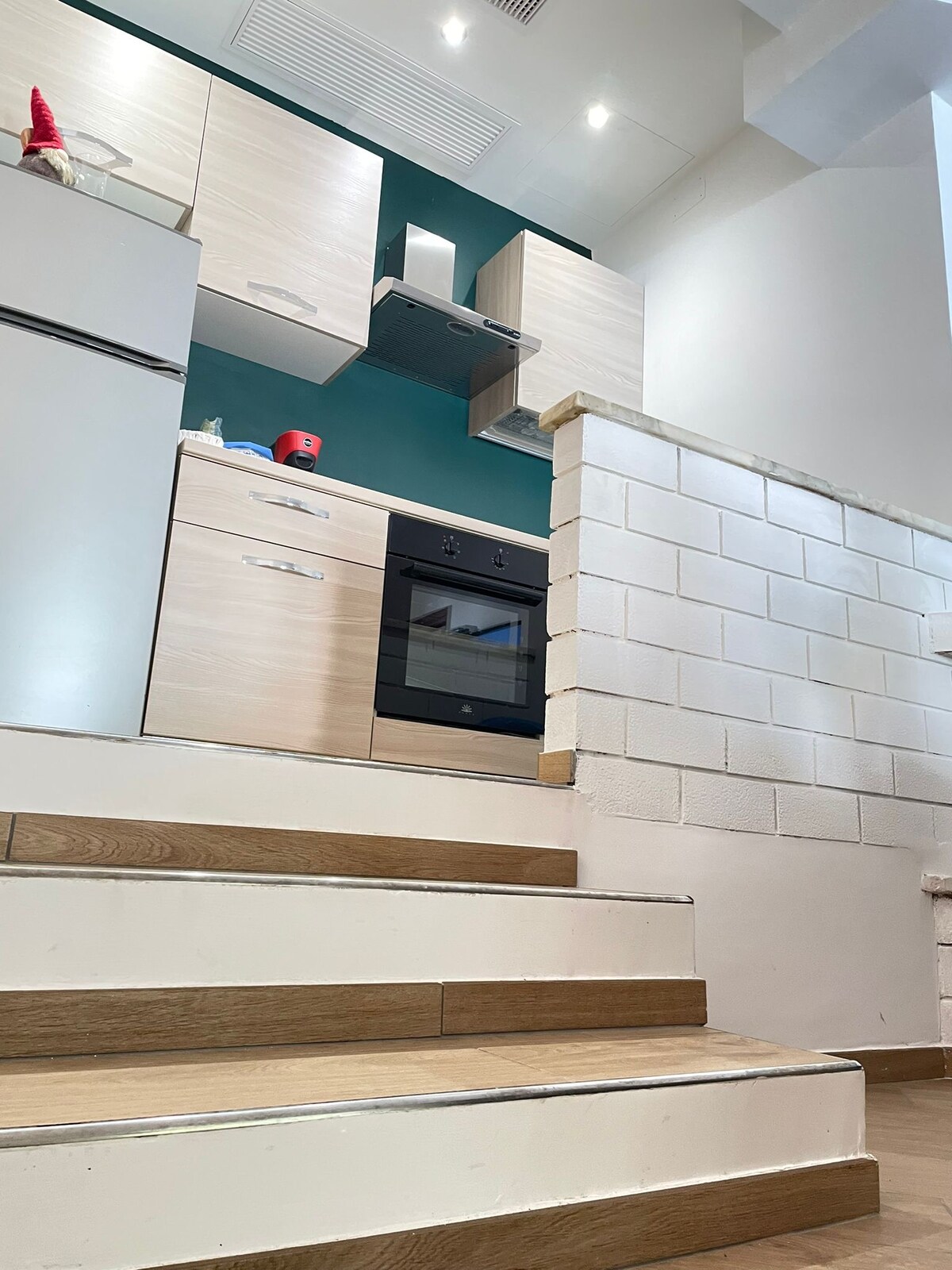 A modern kitchen area is visible at the top of a set of steps, featuring light wood cabinetry and a built-in oven. A contrasting teal wall adds a splash of color. The layout emphasizes functionality with various appliances discreetly integrated.