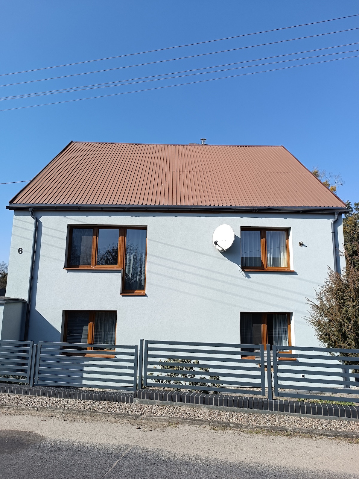 The exterior of a two-story house is presented, featuring a light blue facade and a brown, sloped roof. Large windows with wooden frames are visible, and a satellite dish is mounted on the side. A metal fence surrounds the property, enhancing privacy.