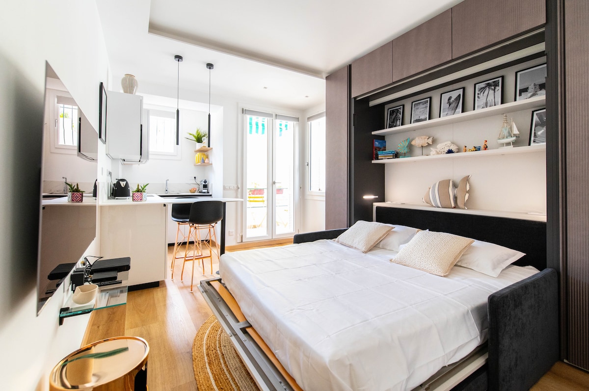 A modern studio apartment features a convertible bed positioned in front of a stylish wall unit. Above the bed, framed photographs are arranged neatly. Large windows allow natural light to fill the space, enhancing the open layout that includes a kitchenette visible to the left.