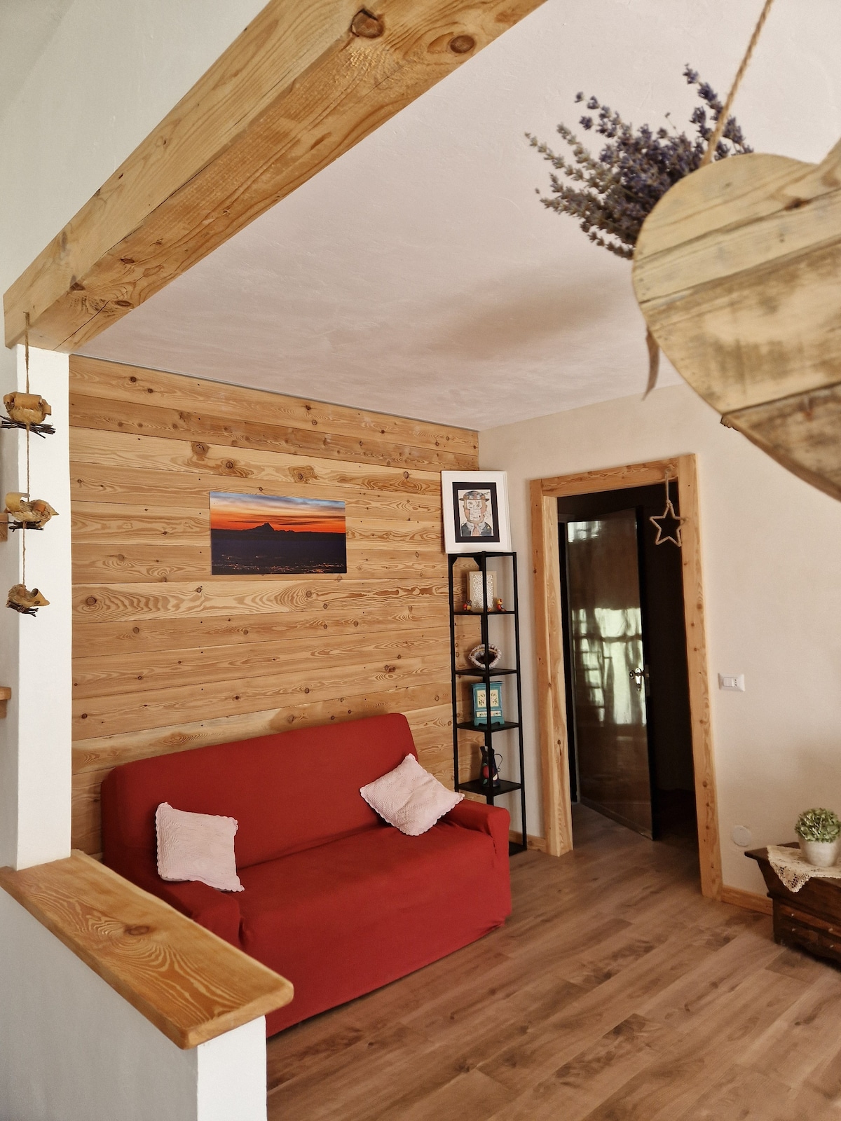 A cozy living area features warm wooden paneling with a piece of artwork on the wall. A red sofa is positioned for comfort, complemented by a bookshelf holding decorative items. A doorway in the background leads to another space, partly concealed by a gentle light.