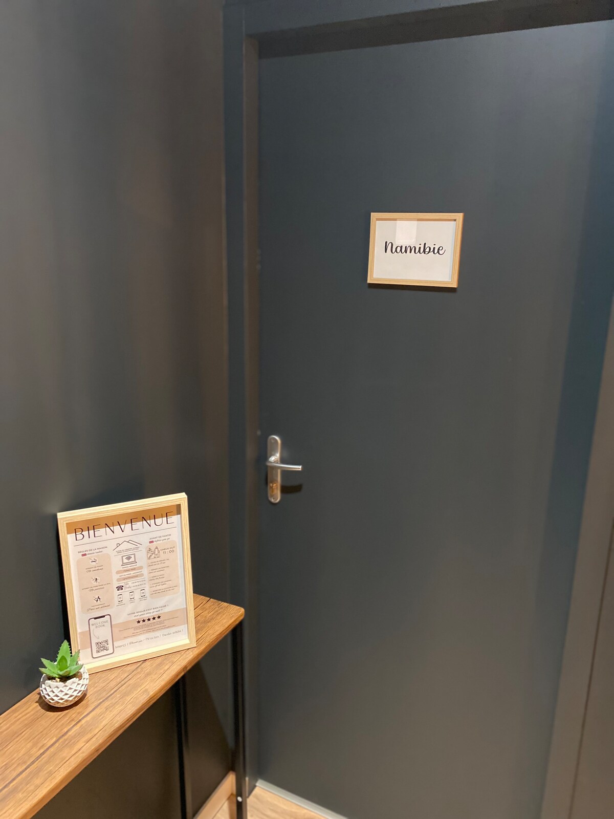A dark blue door labeled 'Namibie' is visible, framed by a wooden console table. A welcome sign and information sheet are displayed on the table, alongside a small decorative plant, creating a warm entrance to the studio.