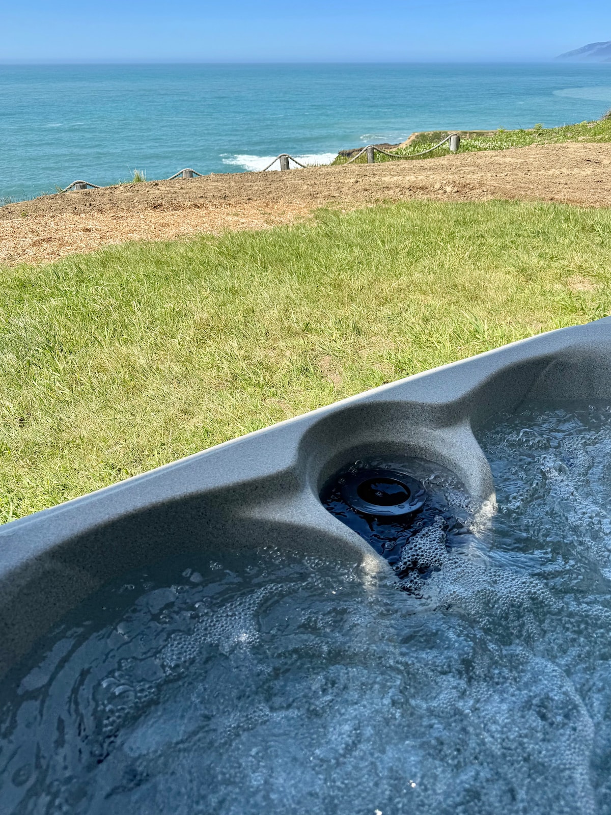 A luxurious jetted tub is positioned on a grassy area, offering a view of the Pacific Ocean. Gentle waves can be seen crashing in the distance, creating a serene backdrop for relaxation.