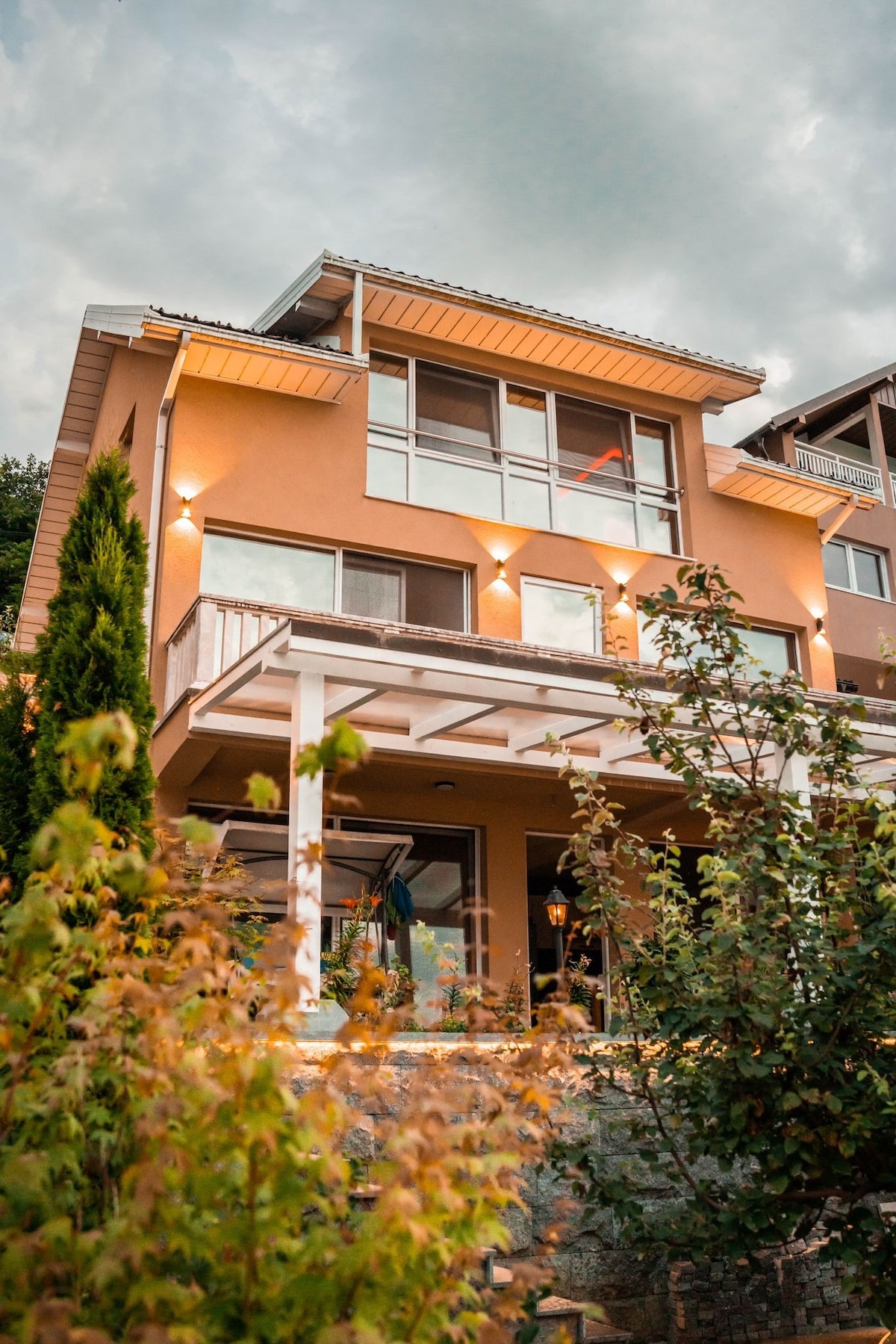 The villa is presented with a charming exterior, featuring multiple levels and large glass windows that enhance natural light. A covered porch is visible in front, surrounded by lush greenery. Soft lighting illuminates the structure, creating a welcoming atmosphere in the evening sky.