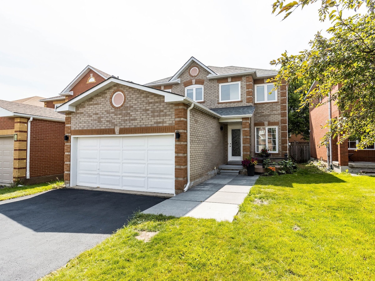 The exterior of a two-story house features a brick facade with soft brown tones. A spacious driveway leads to a double garage. Large windows allow natural light into the home, while a small garden area adds greenery along the side. A tree provides shade to the front yard.