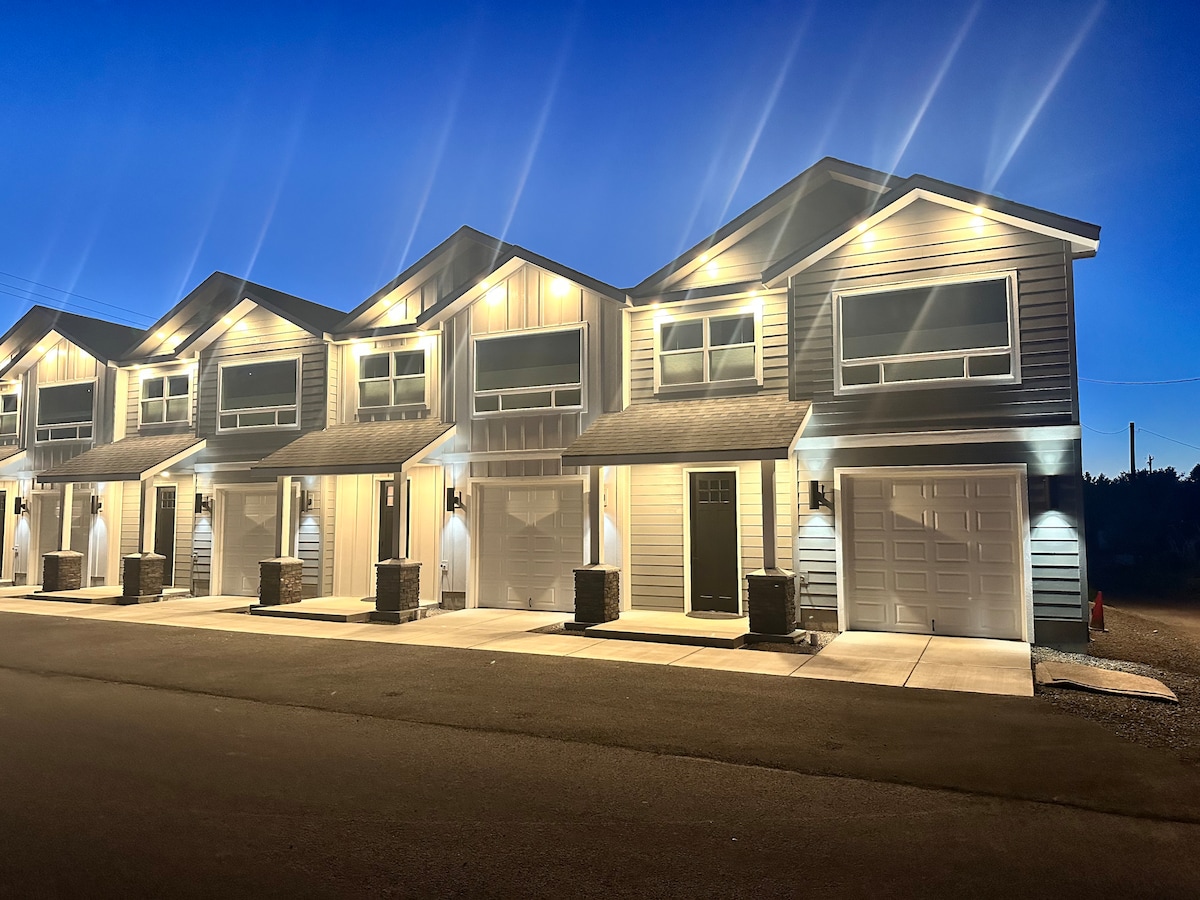 The exterior of the Alder St townhomes is highlighted by warm lighting at dusk, showcasing a row of modern two-story units. Each unit has a welcoming front porch and a single-car garage, with a paved driveway extending in front.
