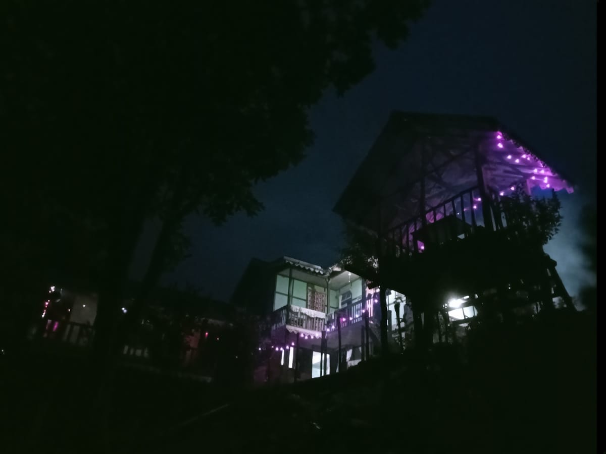 A multi-level building is shown at night, illuminated by soft purple lights. The structure features wooden balconies and large windows, creating an inviting ambiance. Surrounding trees provide a natural backdrop, enhancing the peaceful setting.