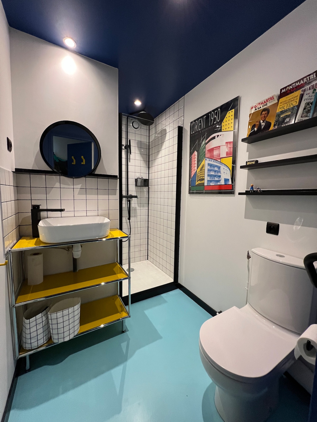 A modern bathroom is shown, featuring a sleek sink mounted on a yellow shelf with open storage. A shower with glass doors is visible, along with a circular mirror. The colorful wall art adds a touch of vibrancy to the tiled walls.