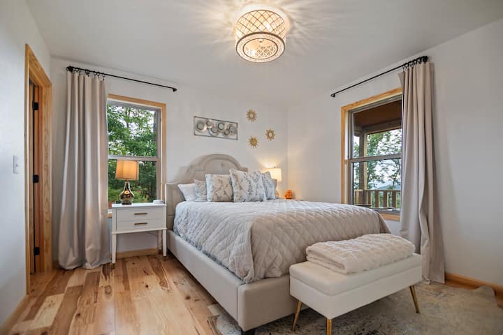 This serene bedroom features a plush queen bedlarge windows with mountain views, light wooden floors, and neutral decor. A stylish chandelier and cozy accents create a tranquil ambiance, perfect for unwinding at the 270 Degree EverLight Escape Cabin.