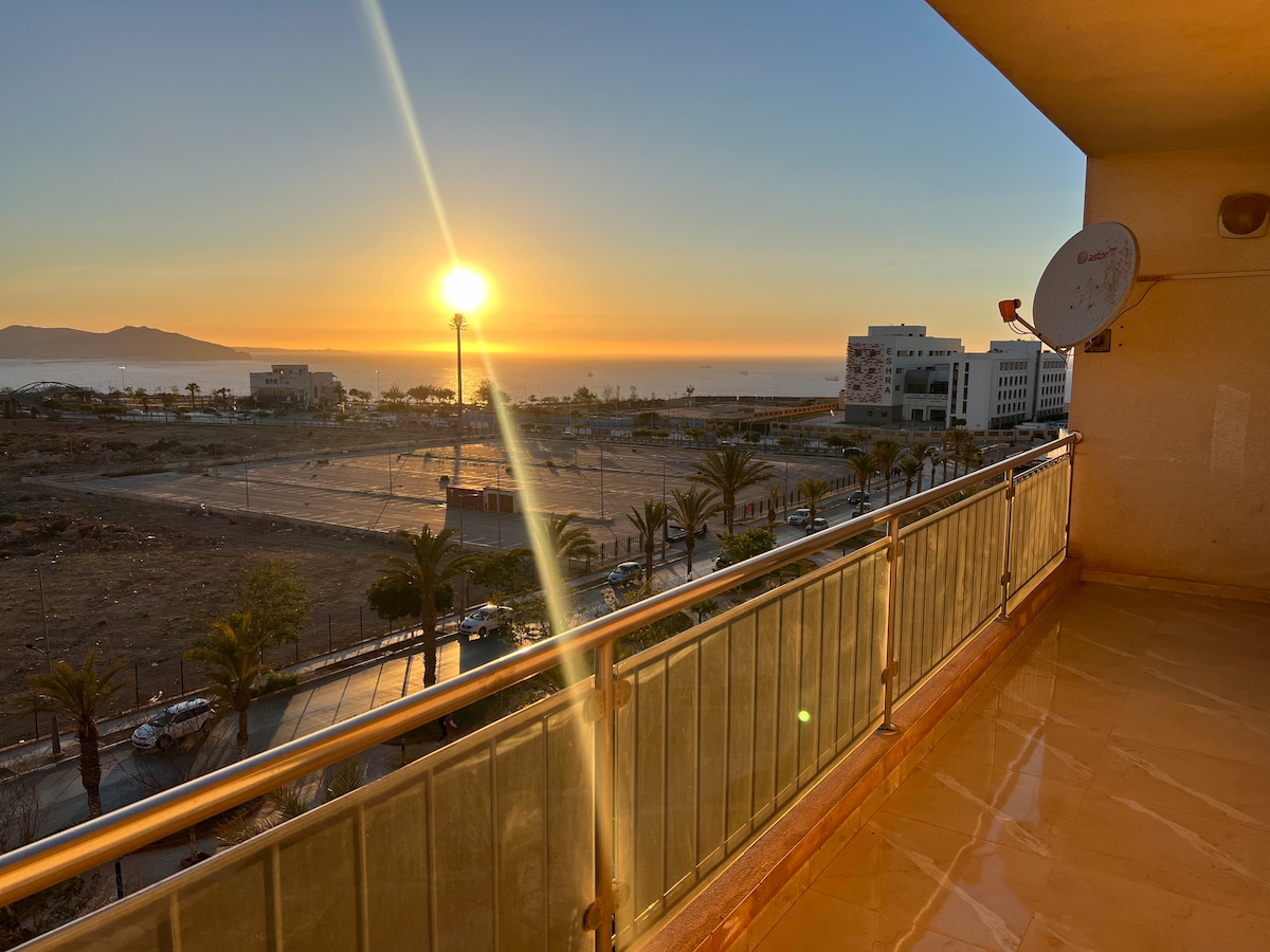 A spacious balcony offers a view of a vibrant sunset over the horizon, where the sun is partially visible. Palm trees align the area below, creating a relaxed atmosphere. The balcony features a glass railing, enhancing the open feeling and connection to the outdoors.