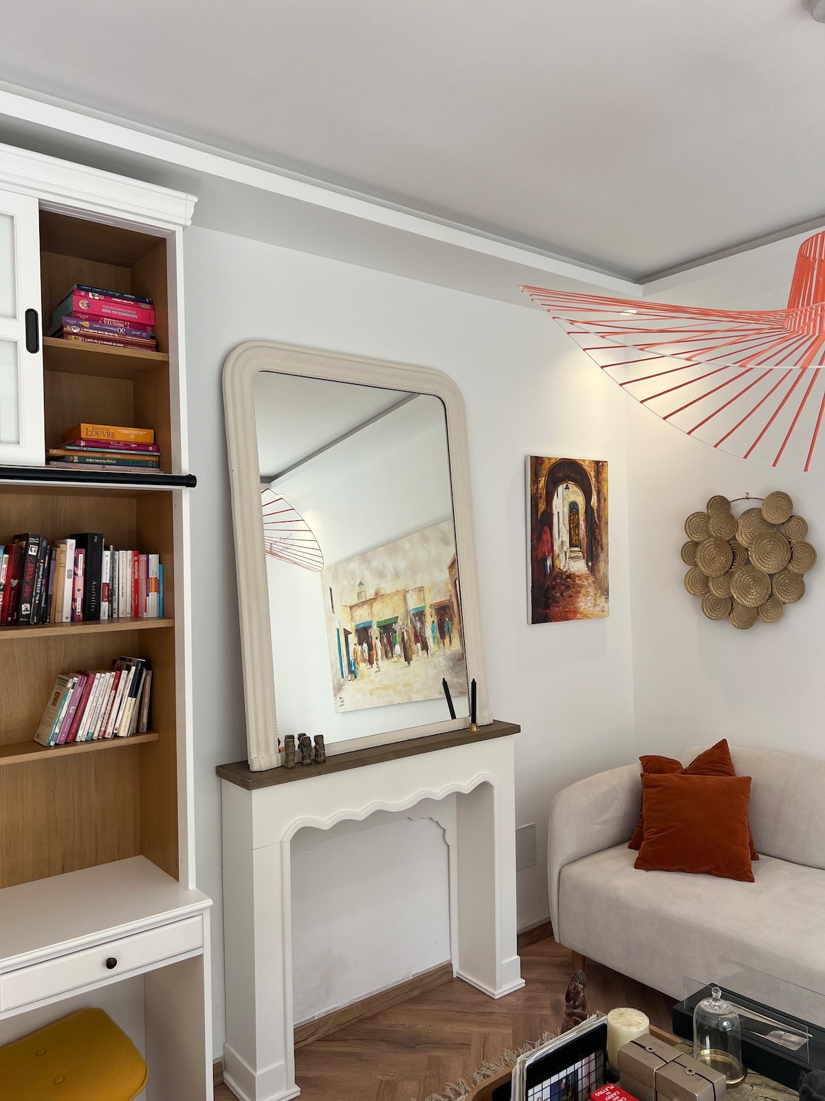 A light-filled lounge area is featured, showcasing a plush sofa with orange accent pillows. A large, decorative mirror reflects a colorful piece of wall art. A bookshelf holds assorted books, while unique wall decorations add character to the inviting space.