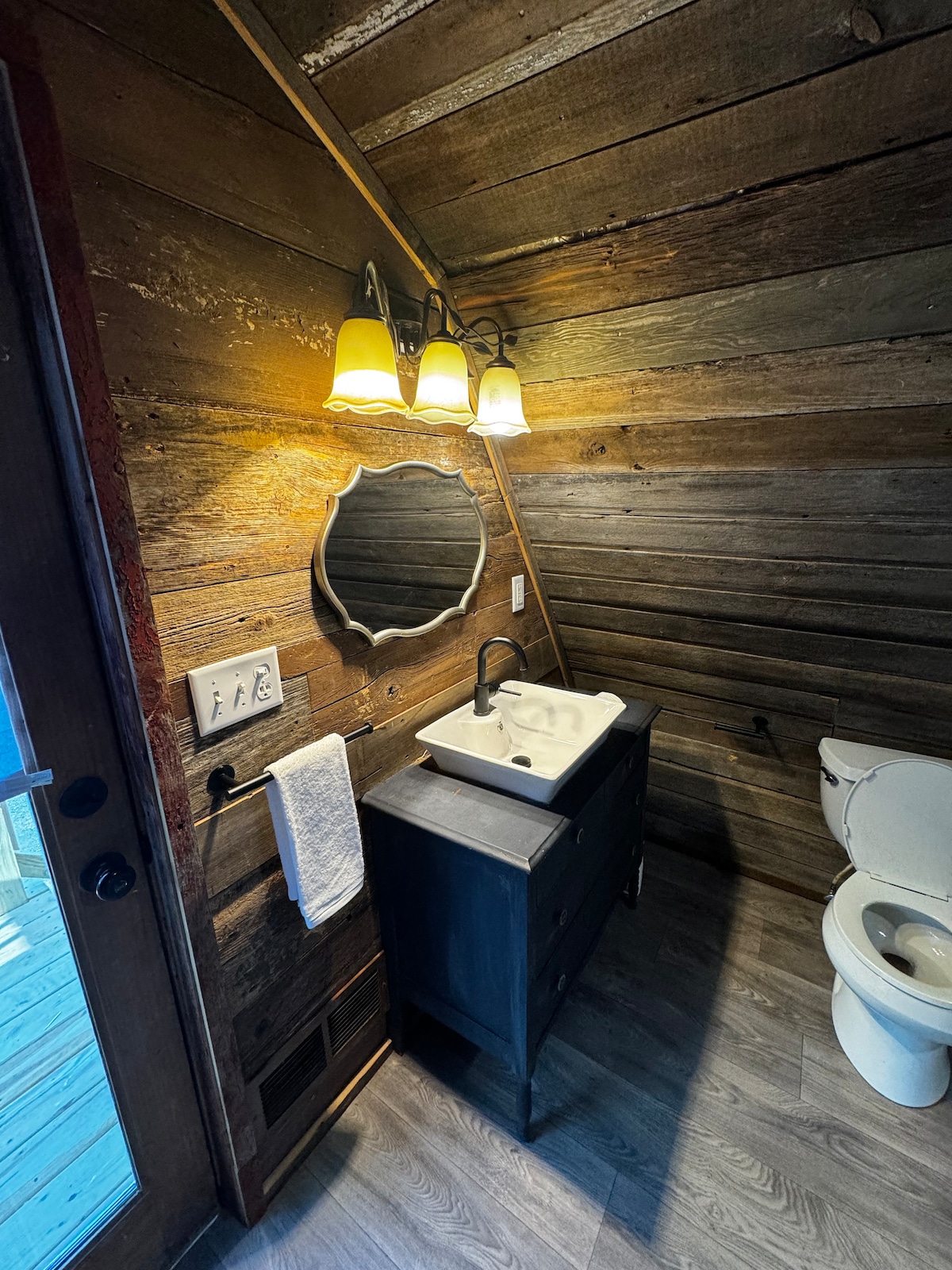 A rustic bathroom features wooden walls and a modern sink atop a dark cabinet. Soft lighting from three lamps reflects off a round mirror. A white towel hangs beside the sink, and a toilet is positioned nearby, enhancing functionality in a warm setting.