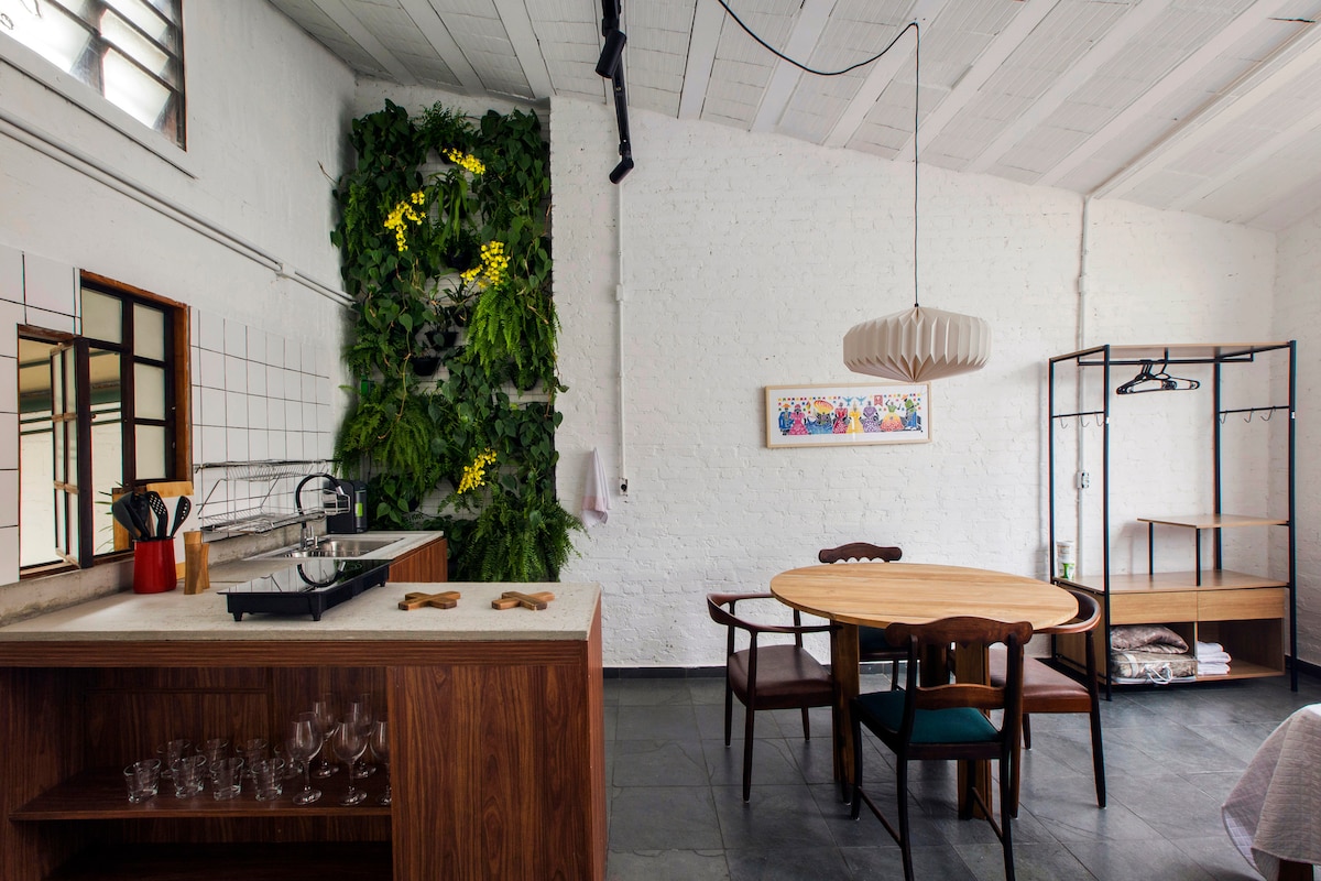 A modern dining area features a round wooden table accompanied by wooden chairs. A vibrant vertical garden adds greenery to the space, while a well-equipped kitchen is positioned along one wall, showcasing stylish cabinetry and counterspace. Natural light filters through a large window.