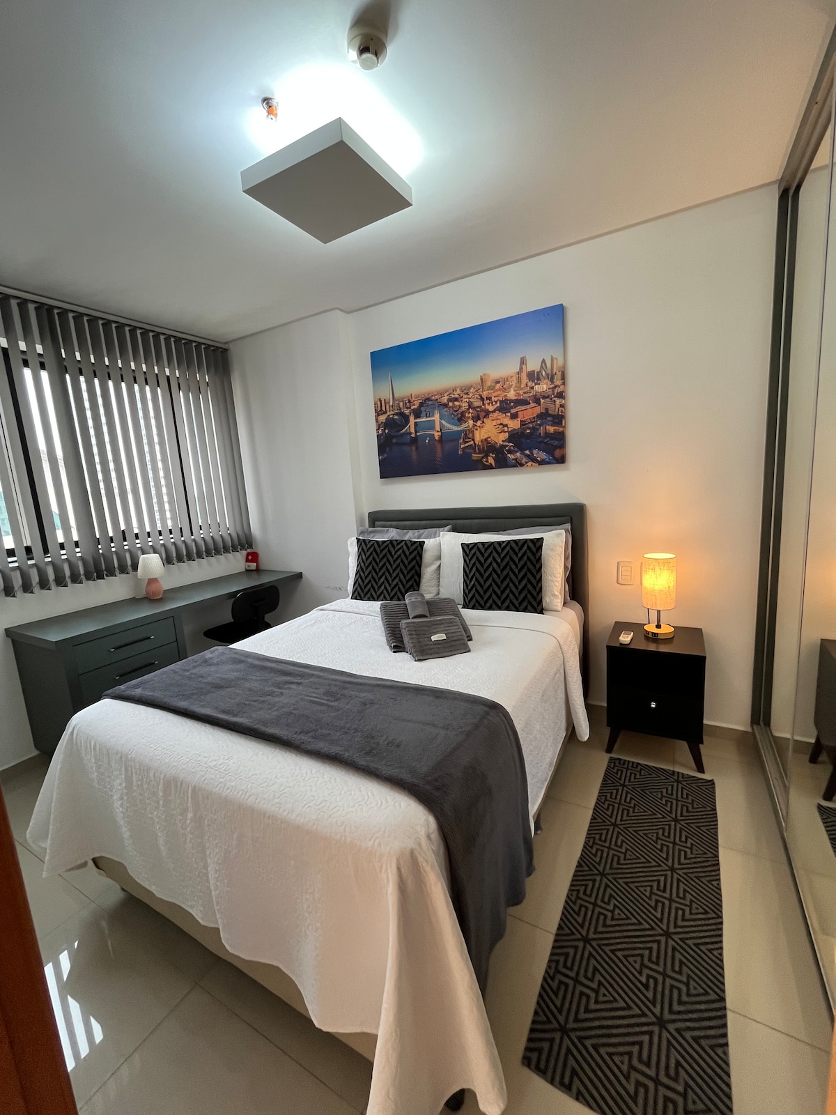 A well-appointed bedroom features a spacious bed adorned with a light blanket and decorative pillows. Soft lighting is provided by a bedside lamp beside a dark nightstand. A large mirror reflects the modern décor, and a cityscape is displayed above the bed, complementing the neutral tones.