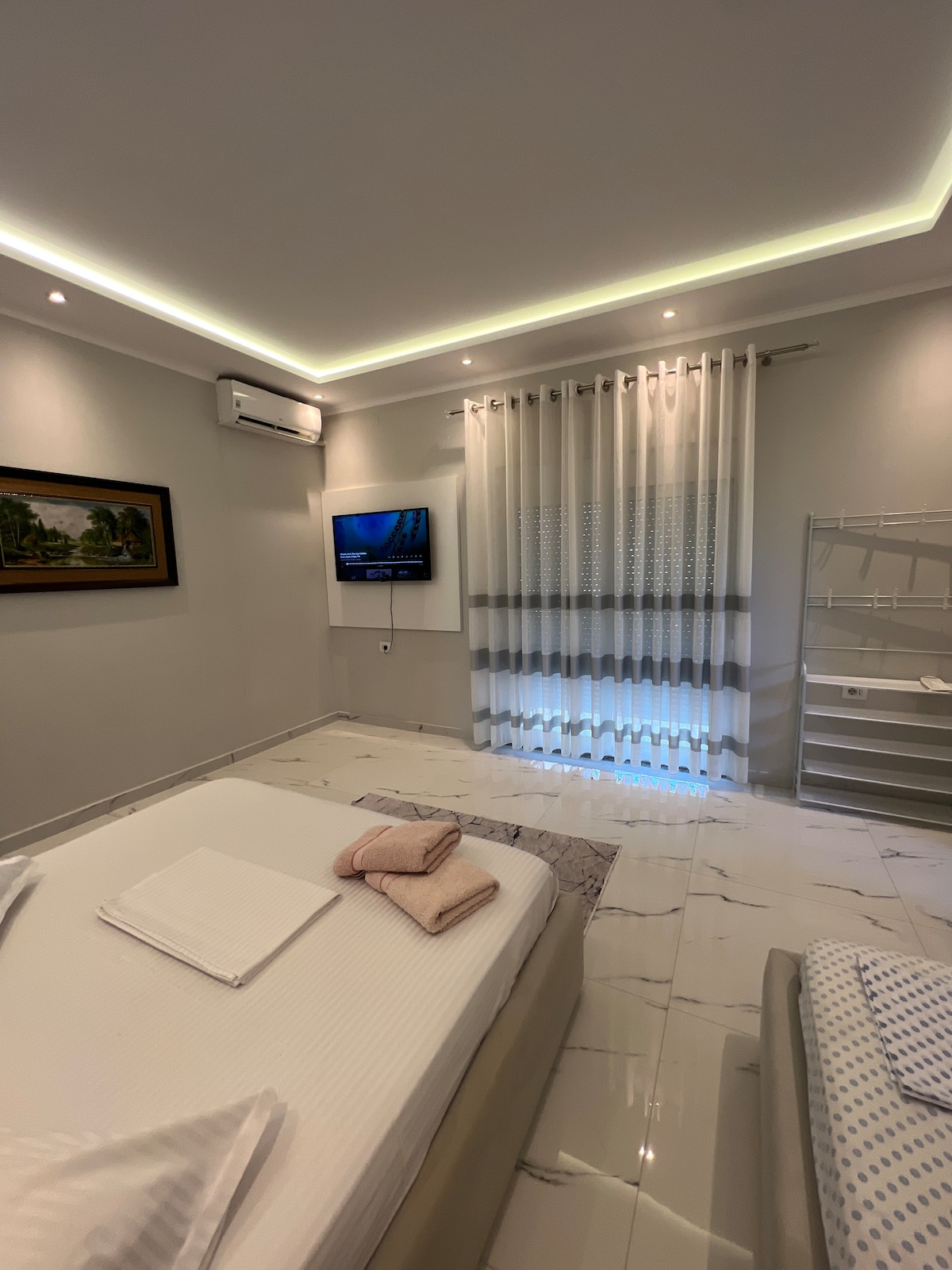 A bright and airy bedroom features a spacious bed with crisp white linens and soft pink towels neatly arranged on top. Natural light filters through sheer white curtains, illuminating the space. A wall-mounted TV is positioned for viewing, and a minimalist clothes rack is visible.