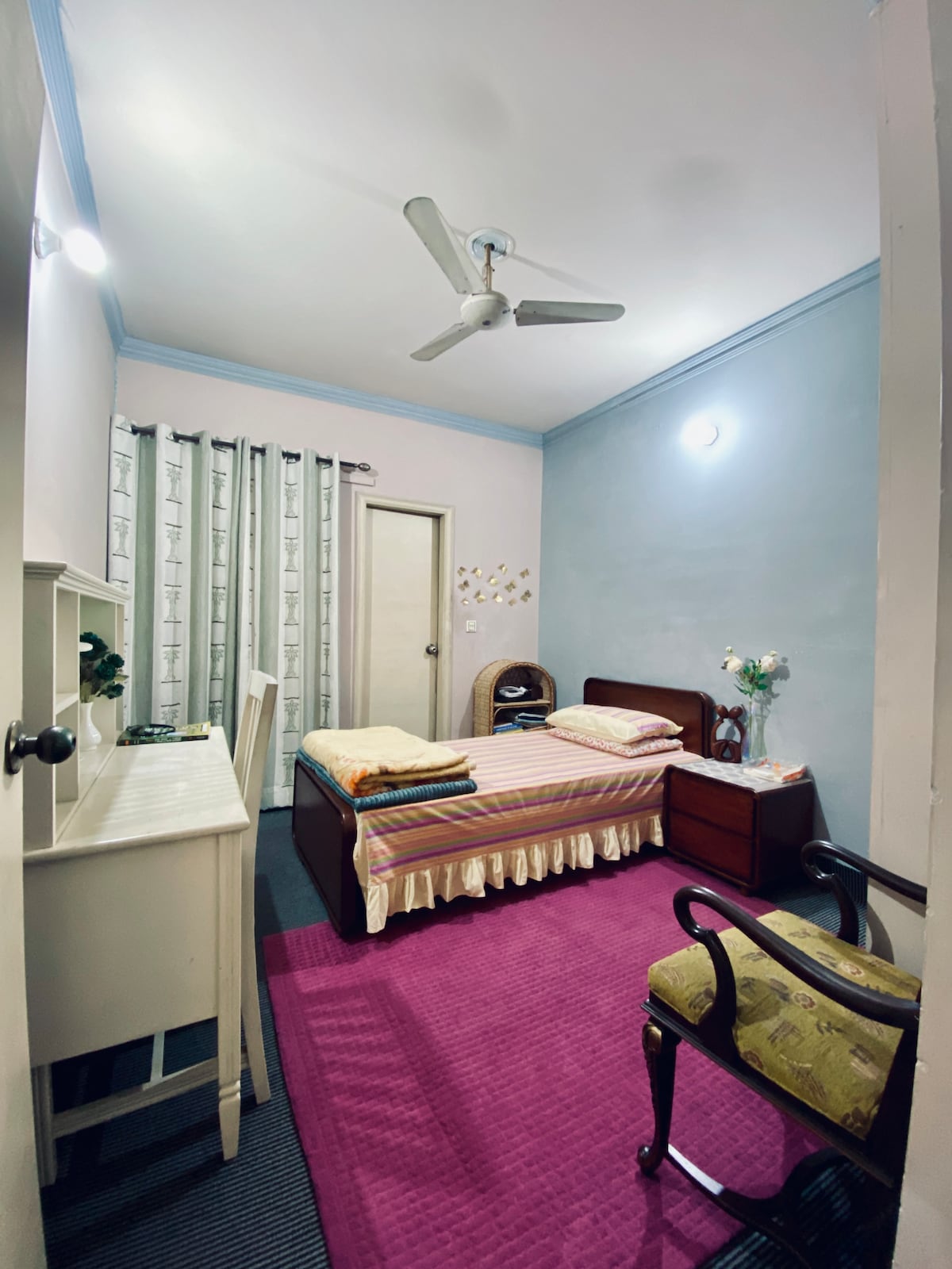 A modest room is presented with a bed covered in light-colored linens, accompanied by a bedside table. A light fixture illuminates the space, while a ceiling fan is visible above. A patterned rug lies on the floor, and a tall window is dressed with light curtains.