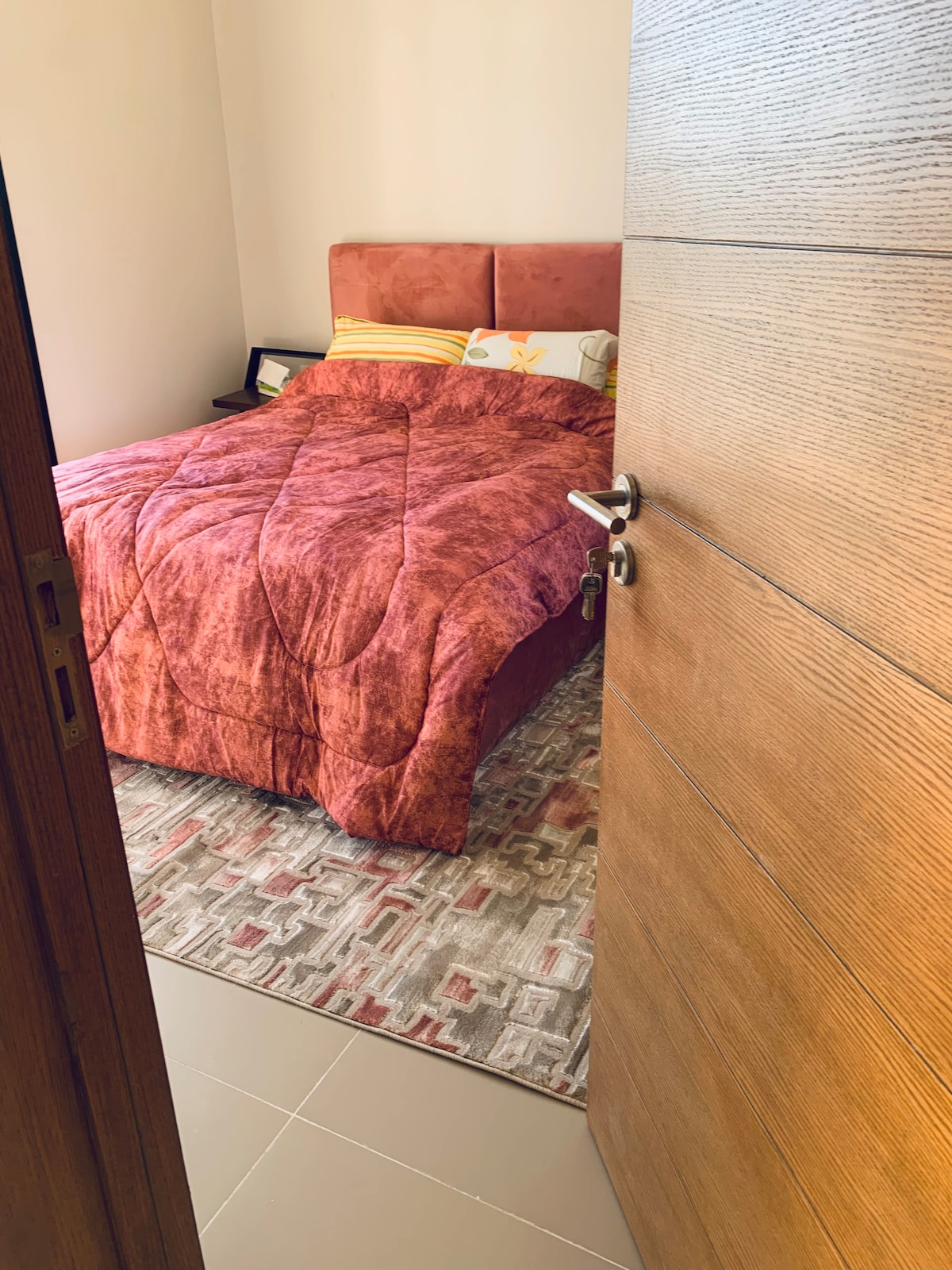 A warm and inviting bedroom is visible through a partially open wooden door. A spacious bed, dressed in a textured maroon comforter, takes center stage. The room features a neutral-colored rug and subtle decorative elements, creating a cozy atmosphere.