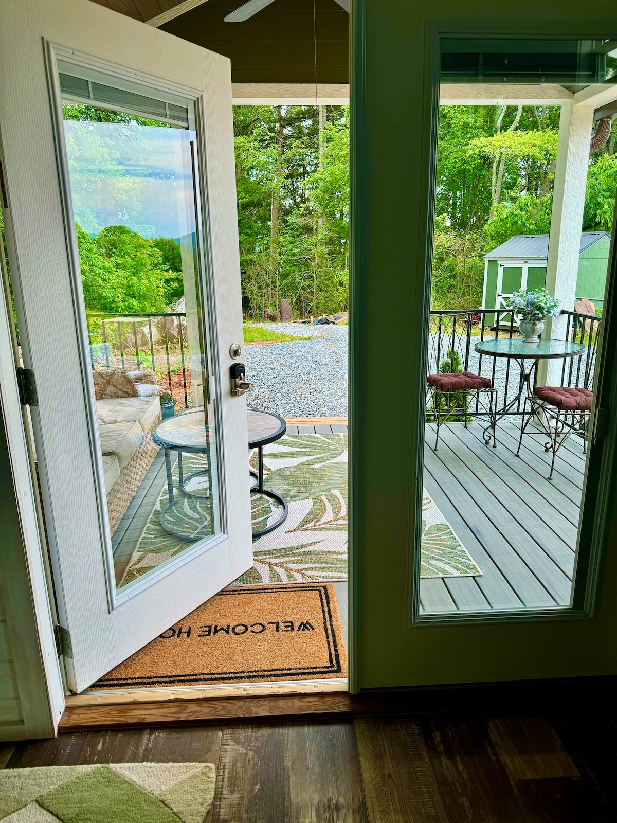 An open door reveals a seating area with a table and two barstools. The outdoor space features a patterned rug and a glimpse of greenery, with a small shed visible in the background. Warm light enhances the inviting atmosphere of the entrance.